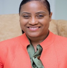 Smiling woman in a coral blazer and olive-green blouse, seated indoors.