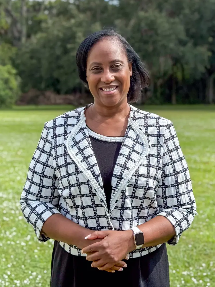 Smiling woman standing outdoors in a green park, wearing a black dress and a white plaid blazer, facing the camera.